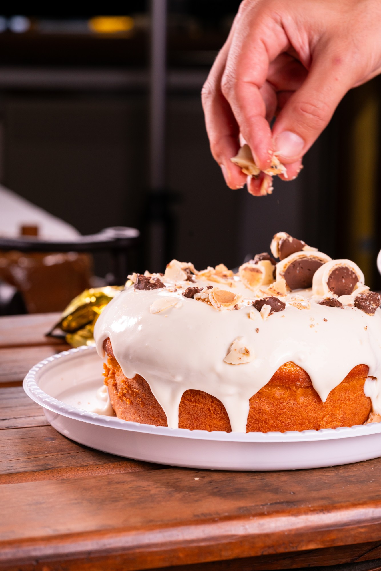 Bolo com cobertura branca sendo finalizado com pedaços de chocolate